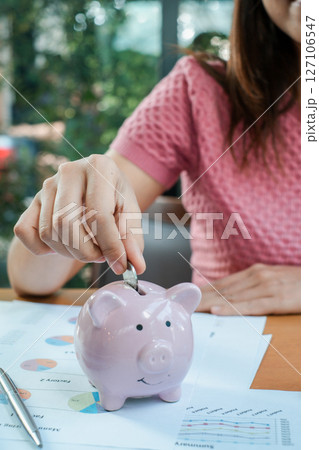 Close-up of a woman placing a coin into a pink piggy bank, surrounded by financial documents, symbolizing savings and financial planning. Close-up of a woman placing a coin into a pink piggy bank, surrounded by financial documents, symbolizing savings and financial planning. 127106547