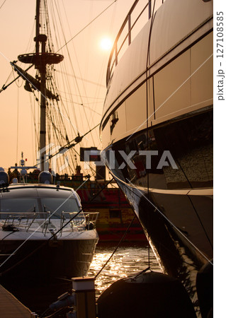 A yacht club at sunset, yachts and sailboats are moored to the berths, huge fenders hang on mega yachts, thick ropes, reflections in the glossy sides of boats, the glare of sun on water surface 127108585