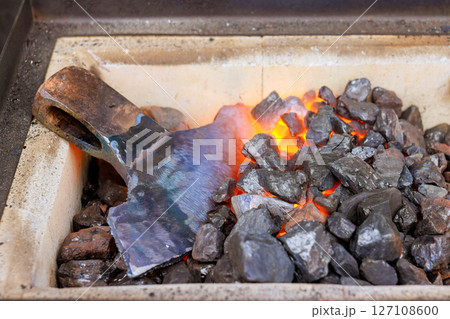 Blacksmith heats large metal tool over glowing coals, during axe making of metalworking outdoors. 127108600