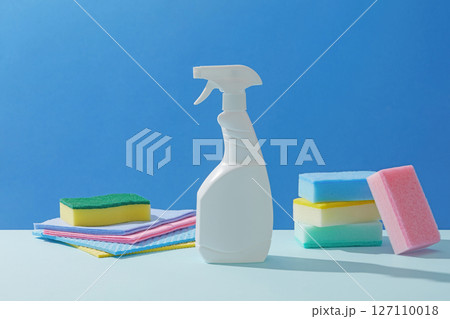 White spray bottle surrounded by soft-colored cleaning sponges, paper, and folded towels, photographed on a crisp, clean blue surface. Concept of cleanliness, cleaning services 127110018