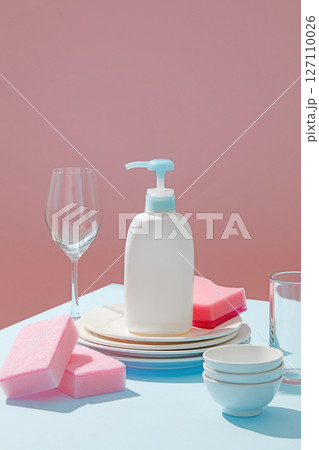 Single soap pump placed among dishes and wine glasses with shadows, standing out against a bright, pastel pink and blue-toned background. Ad template or package design for cleaning product 127110026