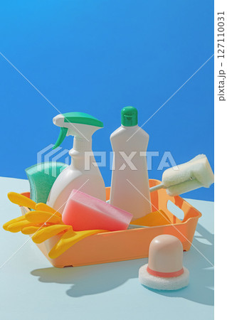 Bright and playful photo showing plastic cleaning bottles and tools inside a yellow basket on a blue backdrop, ideal for advertising cleaning products. Product bottle unlabeled, blank space for design 127110031