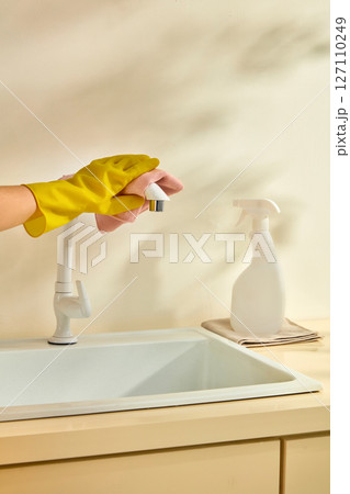 Hands in yellow gloves wiping kitchen counters with spray bottle and sponge. Minimalist and clean living scene in a light-toned kitchen. Empty spray bottle label for design 127110249