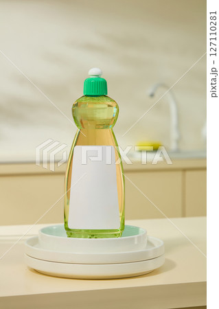 A bottle of detergent with a green cap is placed on a white plate, displayed under soft lighting on a warm-toned kitchen background. Mockup for advertising dishwashing products, label design 127110281