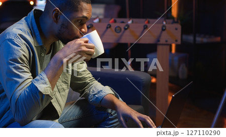 African American young adult working remotely on a laptop under loft neon lights, sitting on a couch in his cozy apartment. Relaxed person in a stylish home highlights modern productivity. Camera B. 127110290