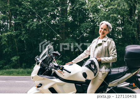 Stylish senior woman with gray hair sitting on motorcycle holding helmet near forest road Stylish senior woman with gray hair sitting on motorcycle holding helmet near forest road 127110438
