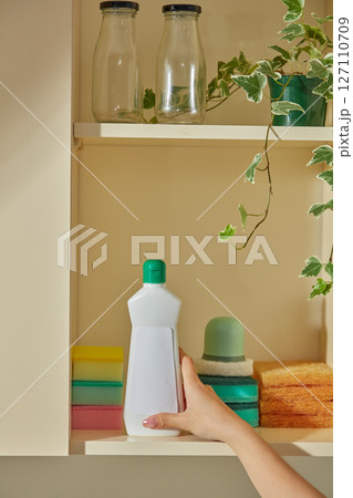 A person places a green dishwashing liquid bottle on a kitchen shelf with neatly arranged jars and a plant, blending cleaning supplies with decor. White bottle without label for design A person places a green dishwashing liquid bottle on a kitchen shelf with neatly arranged jars and a plant, blending cleaning supplies with decor. White bottle without label for design 127110709