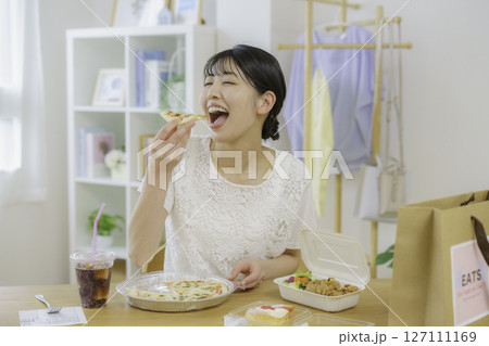 デリバリーで頼んだ食べ物を置いた食卓の前でピザを食べようとする若い女性 127111169