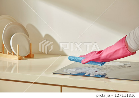 Gloved hands cleaning the stove area with a blue sponge near stacked dishes, highlighting daily kitchen maintenance and home care essentials. Gloved hands cleaning the stove area with a blue sponge near stacked dishes, highlighting daily kitchen maintenance and home care essentials. 127111816