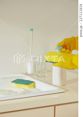 A sponge and pump cleaner placed next to the faucet on a counter, gloved hands preparing for a wipe-down session in a spotless beige kitchen. A sponge and pump cleaner placed next to the faucet on a counter, gloved hands preparing for a wipe-down session in a spotless beige kitchen. 127111819