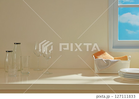 Sunlight spilling onto a countertop with stacked dishes, glassware, and a storage basket, setting a calm mood in a well-kept kitchen. Blank space for display your product 127111833
