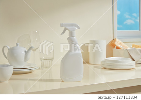 White spray bottle next to clean white teapots and mugs, arranged simply in a bright kitchen with blue sky in the background. 127111834