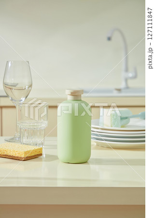 Green bottle and dish sponge beside crystal glasses on a beige surface, blending utility and elegance in a clean kitchen layout. 127111847