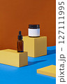 On an orange and blue background, yellow geometric podiums displayed with amber serum bottle and amber jar. Blank space for design label, front view. 127111995