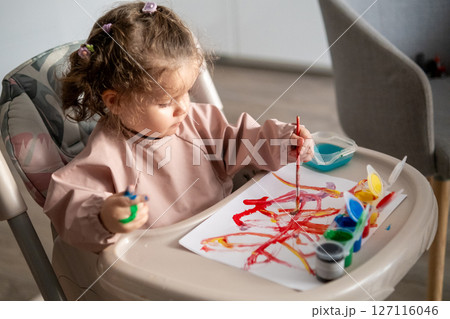 Cute toddler girl painting with gouache at a table near a sunlit window. Natural light highlights her artwork and the joyful atmosphere of creativity. 127116046