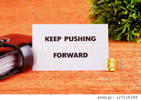 Concept text KEEP PUSHING FORWARD written on a card on a bright orange table next to a black notebook and a small green plant 127116394