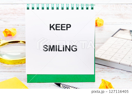 KEEP SMILING written on a desktop notebook with decorative elements such as a calculator, a magnifying glass and yellow origami KEEP SMILING written on a desktop notebook with decorative elements such as a calculator, a magnifying glass and yellow origami 127116405