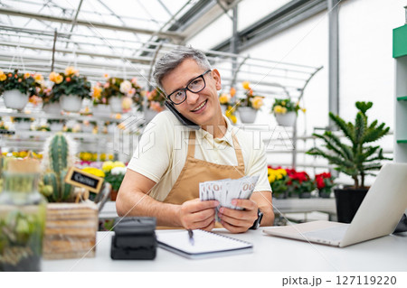 A Caucasian florist, happy with money in hand, smiles while on a phone call in his shop, surrounded by plants and flowers. 127119220