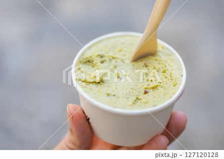 Hand holding cup of pistachio gelato with wooden spoon close-up. Summer dessert, street food, and refreshing sweet treat outdoors. 127120128