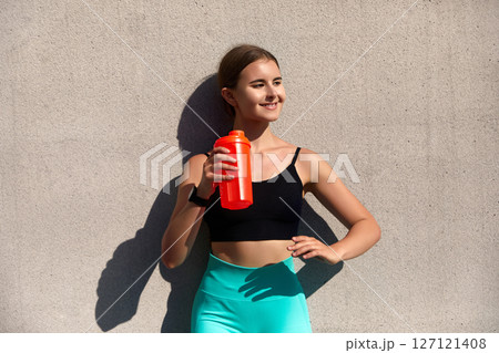 Young woman in activewear holding a bright red water bottle against a concrete wall, showcasing fitness and hydration lifestyle, healthy living concept Young woman in activewear holding a bright red water bottle against a concrete wall, showcasing fitness and hydration lifestyle, healthy living concept 127121408