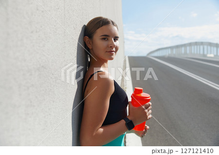 Young woman resting against wall with sports drink bottle on sunny day, showcasing fitness lifestyle and healthy hydration concept Young woman resting against wall with sports drink bottle on sunny day, showcasing fitness lifestyle and healthy hydration concept 127121410