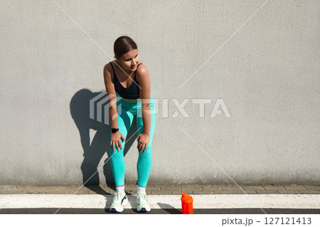 Athletic woman resting after workout against a wall, wearing stylish sportswear, focused on hydration with a water bottle, fitness lifestyle concept Athletic woman resting after workout against a wall, wearing stylish sportswear, focused on hydration with a water bottle, fitness lifestyle concept 127121413