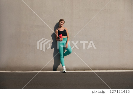 Sports woman in active wear standing outdoors against concrete wall with water bottle and smiling at camera. Fitness girl having a break after workout run. Sports woman in active wear standing outdoors against concrete wall with water bottle and smiling at camera. Fitness girl having a break after workout run. 127121416