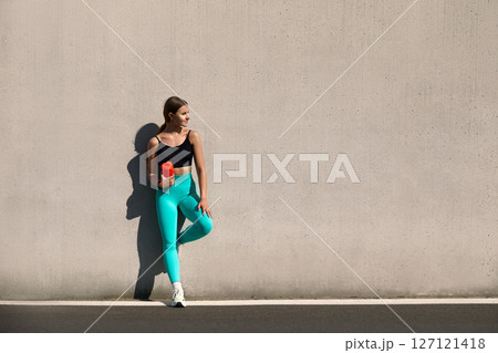 Fitness woman in trendy activewear leans against a gray wall while holding a red dumbbell, embodying a commitment to a healthy lifestyle 127121418