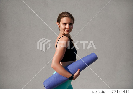 Young sportive woman with a toned physique holds a purple yoga mat while enjoying outdoor exercise. She radiates happiness and embodies a healthy lifestyle during her fitness routine Young sportive woman with a toned physique holds a purple yoga mat while enjoying outdoor exercise. She radiates happiness and embodies a healthy lifestyle during her fitness routine 127121436