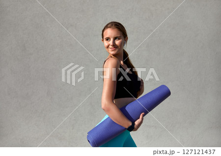 Young woman in athletic wear holding a yoga mat, smiling confidently against a textured gray background, embodying fitness and wellness while preparing for a yoga session or workout routine Young woman in athletic wear holding a yoga mat, smiling confidently against a textured gray background, embodying fitness and wellness while preparing for a yoga session or workout routine 127121437