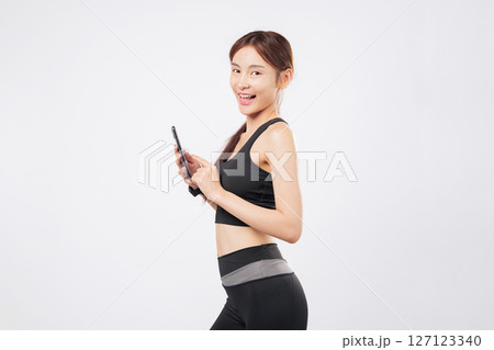 Phone in hand and another phone swipe later, this fit sporty woman stands confidently in workout clothes, showing off her gym dedication and mobile lifestyle. isolated studio white background Phone in hand and another phone swipe later, this fit sporty woman stands confidently in workout clothes, showing off her gym dedication and mobile lifestyle. isolated studio white background 127123340