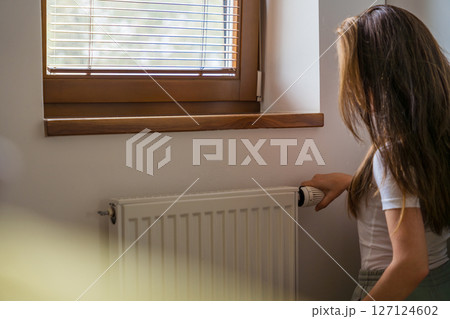 A woman with long dark hair adjusts the radiator in a cozy home as the heating season begins. 127124602