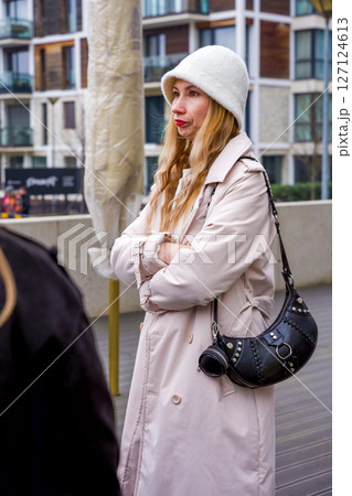 Elegant woman in a white bucket hat and beige trench coat in a modern cityscape Elegant woman in a white bucket hat and beige trench coat in a modern cityscape 127124613