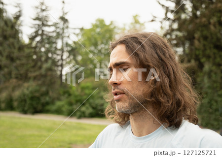 Young caucasian man with long hair walks outdoors at forest in public park. He feel depression and sad. Close up. Outdoors 127125721