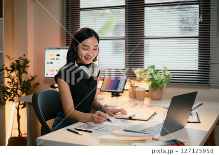 Graphic designer woman is sitting at a desk with a laptop and a notebook. She is wearing headphones and smiling Graphic designer woman is sitting at a desk with a laptop and a notebook. She is wearing headphones and smiling 127125958