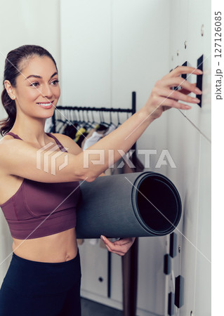 Smiling young woman with a rolled mat opening a locker in the gym 127126085