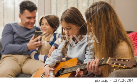 Young girl learning to play guitar with family at home Young girl learning to play guitar with family at home 127126294