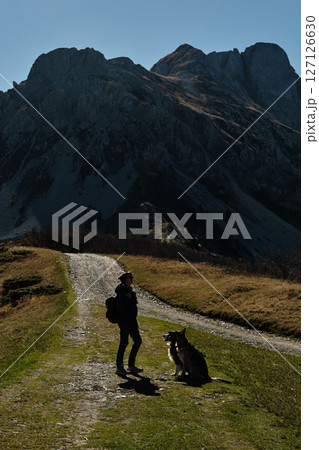 Woman hiking with two dogs - German and Australian shepherds on a mountain trail in the Komovi range, Montenegro, surrounded by dramatic peaks and golden autumn meadows. Hiking with pet concept Woman hiking with two dogs - German and Australian shepherds on a mountain trail in the Komovi range, Montenegro, surrounded by dramatic peaks and golden autumn meadows. Hiking with pet concept 127126630