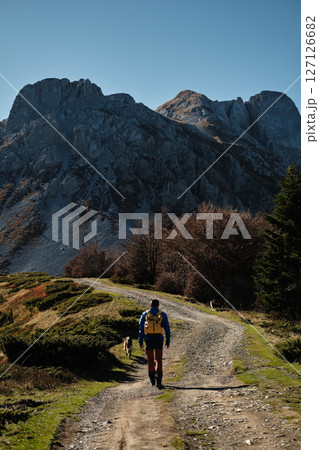 Male solo hiker with a yellow backpack walks along a winding dirt trail in the Komovi mountains in Montenegro, accompanied by two dogs on a sunny autumn day. Rear view. Hiking with pet concept Male solo hiker with a yellow backpack walks along a winding dirt trail in the Komovi mountains in Montenegro, accompanied by two dogs on a sunny autumn day. Rear view. Hiking with pet concept 127126682