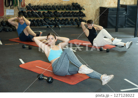 Concentrated young fitness woman doing abdominal exercise on yoga mat during group workout in gym. CrossFit healthy concept. 127128022