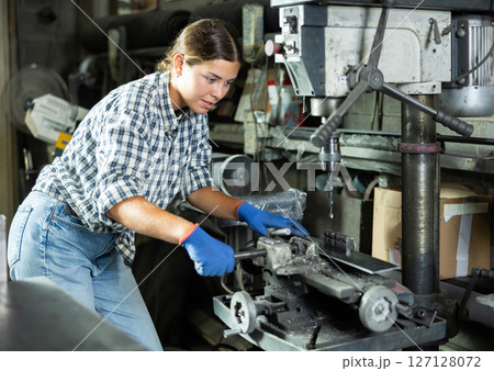 Girl worker at metallurgical plant drills hole in metal plate using drilling machine 127128072