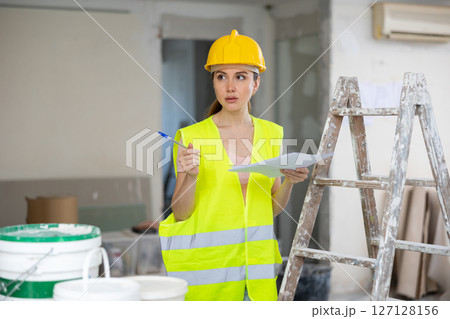 Woman foreman in a protective helmet and yellow vest checks the completed construction work on drawing 127128156