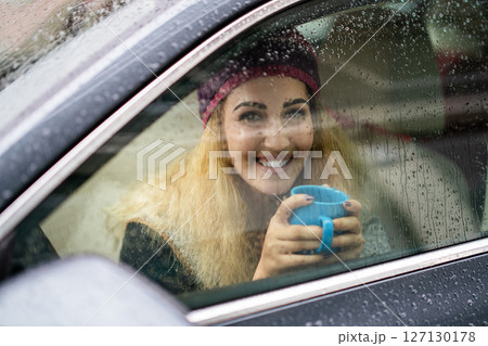 Smiling young blond woman holding coffee in the car on rainy day. 127130178
