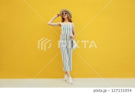 Summer outfit, stylish beautiful young woman posing in white striped jumpsuit against yellow wall 127131054