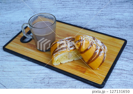 A sliced mocha golden loaf with chocolate drizzle and powdered sugar, served with a glass of hot chocolate on a wooden board. 127131650