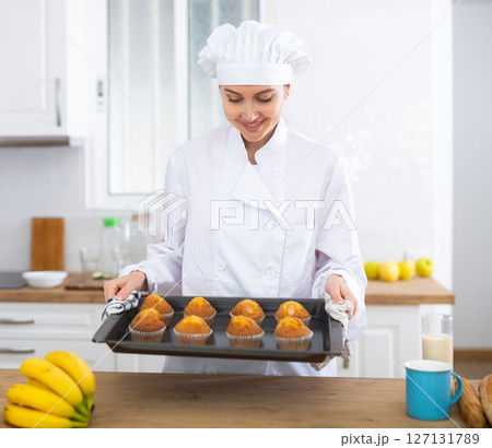 Proffesional woman cook in white uniform holding sheet pan with just baked cupcakes 127131789