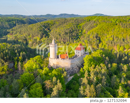 Kokorin Castle stands majestically amidst lush greenery in Czechia during a sunny evening. The aerial view showcases its ancient architecture surrounded by rolling hills and vibrant trees. 127132094