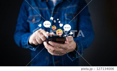 A person in a denim jacket interacts with a smartphone, surrounded by floating emojis, chat bubbles, hearts, and icons representing social engagement and network connectivity in the digital world. 127134006