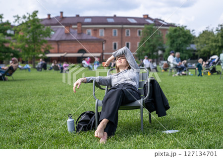 Tired woman stretched on chair in city park blocking sunlight with hand, dozing with drink nearby 127134701