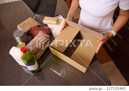 Person Packing Essential Food Items into a Cardboard Box on a Table with Supplies 127135465